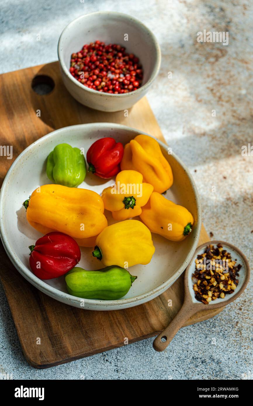 Gros plan d'un bol de piments rouges, jaunes et verts bonnet écossais sur une planche à découper avec des flocons de piment et des grains de poivre rose Banque D'Images