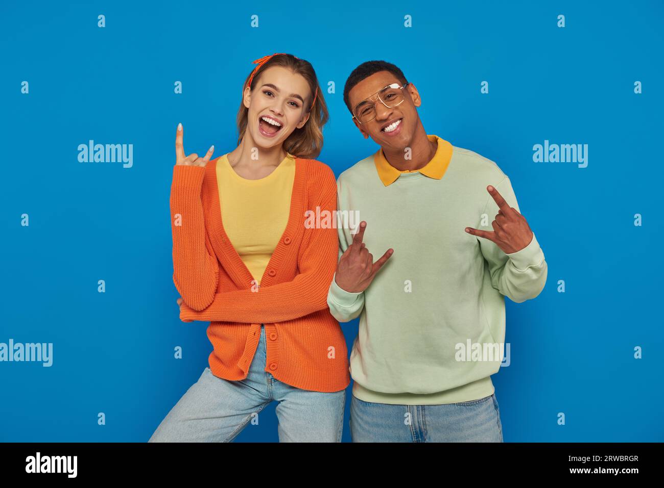 excité et jeune couple interracial en tenue décontractée gestuant sur fond bleu, montrant signe de roche Banque D'Images