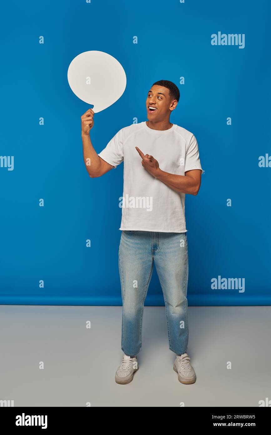 homme afro-américain excité en jean denim pointant vers la bulle de discours sur fond bleu, pleine longueur Banque D'Images