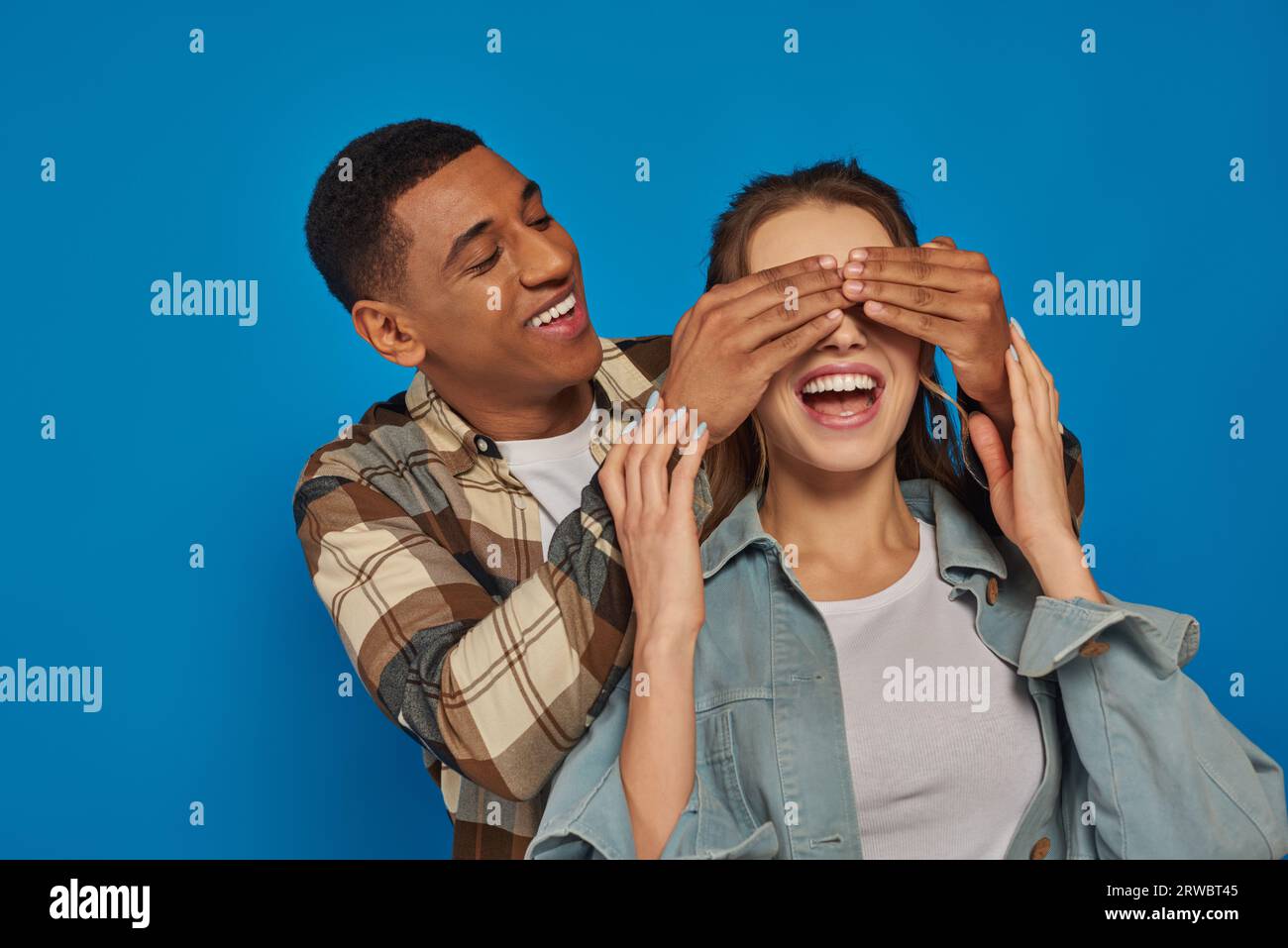 homme afro-américain excité couvrant les yeux de la femme avec la bouche ouverte sur fond bleu, peekaboo Banque D'Images