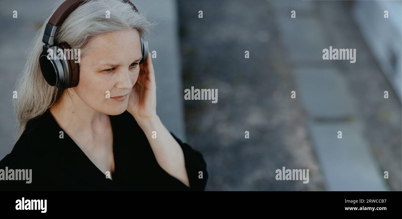 Vue de la tête d'une belle femme aux cheveux gris assise sur des escaliers en béton en ville, écoutant de la musique à travers des écouteurs sans fil. Banque D'Images