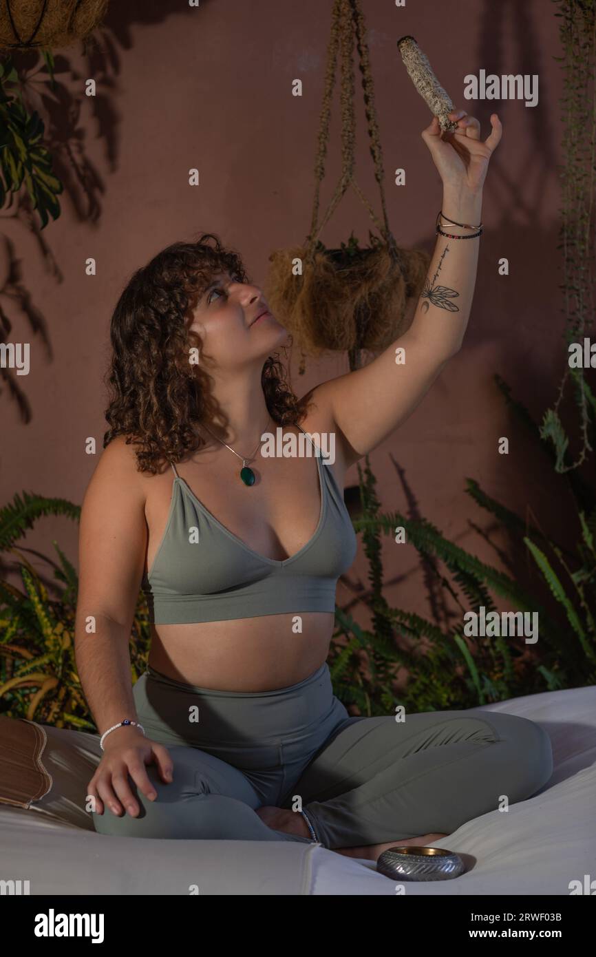 Portrait d'une jeune femme aux cheveux noirs bouclés, assise jambes croisées en méditation tenant un bâton de fumage avec un bras levé Banque D'Images