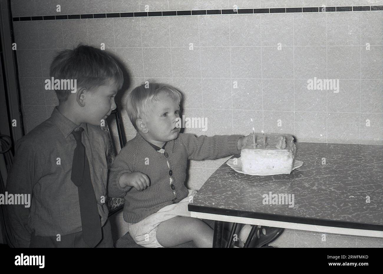 Années 1960, joyeux anniversaire, jeune écolier debout par un frère de deux ans avec un petit gâteau assis sur le dessus de table formica, Angleterre, Royaume-Uni, bougies allumées ?? Que dois-je faire ? Banque D'Images