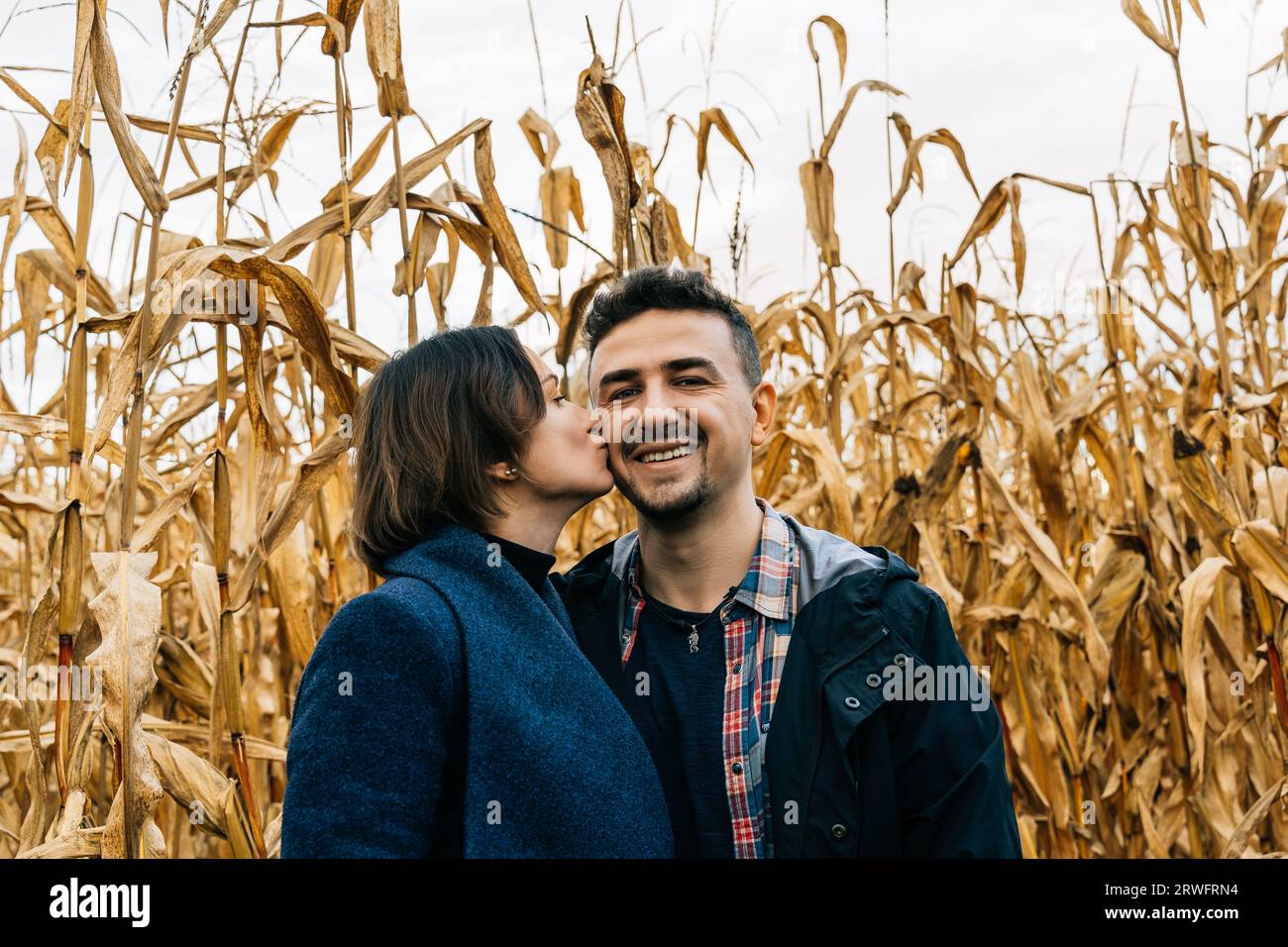 Femme embrassant l'homme souriant heureux sur la joue debout en automne dans un champ de maïs Banque D'Images