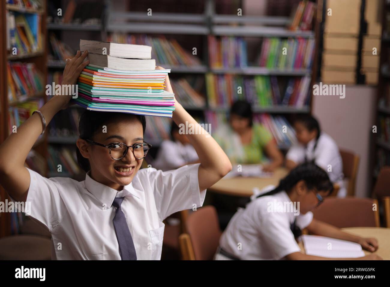 Heureuse adolescente étudiante tenant des livres sur sa tête, debout dans la bibliothèque, travail acharné, préparation aux examens, pression, éduquer, stresser. Banque D'Images