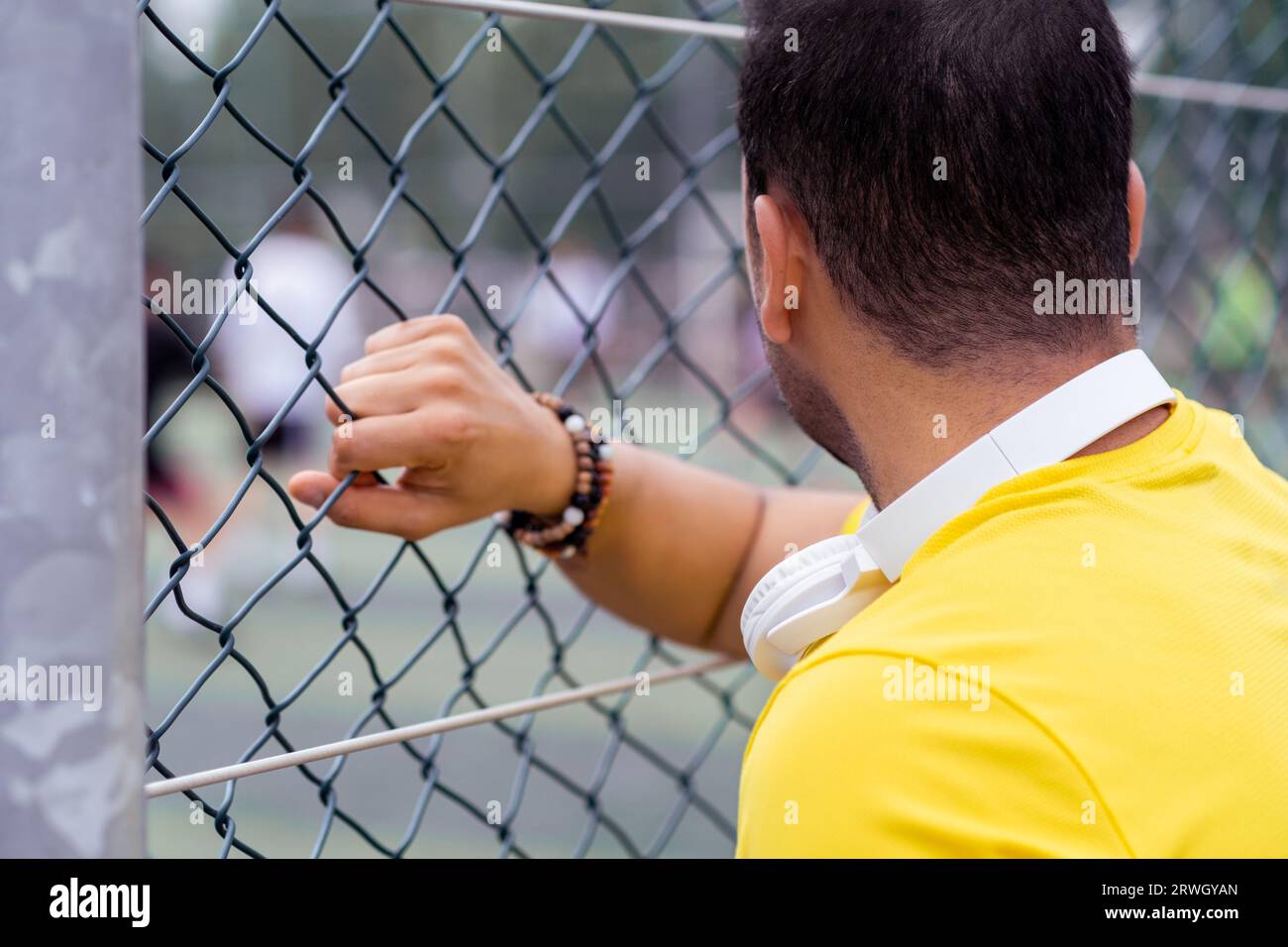 homme avec écouteurs regarde les joueurs de football sur l'arène urbaine, regardant à travers la grille de clôture métallique. Banque D'Images