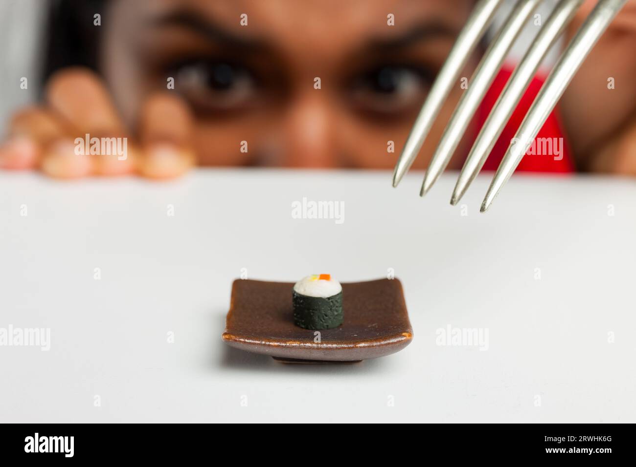Au premier plan : un micro sushi Hosomaki et une fourchette surdimensionnée. La toile de fond floue met en valeur le regard d'une indienne dicté par la faim Banque D'Images