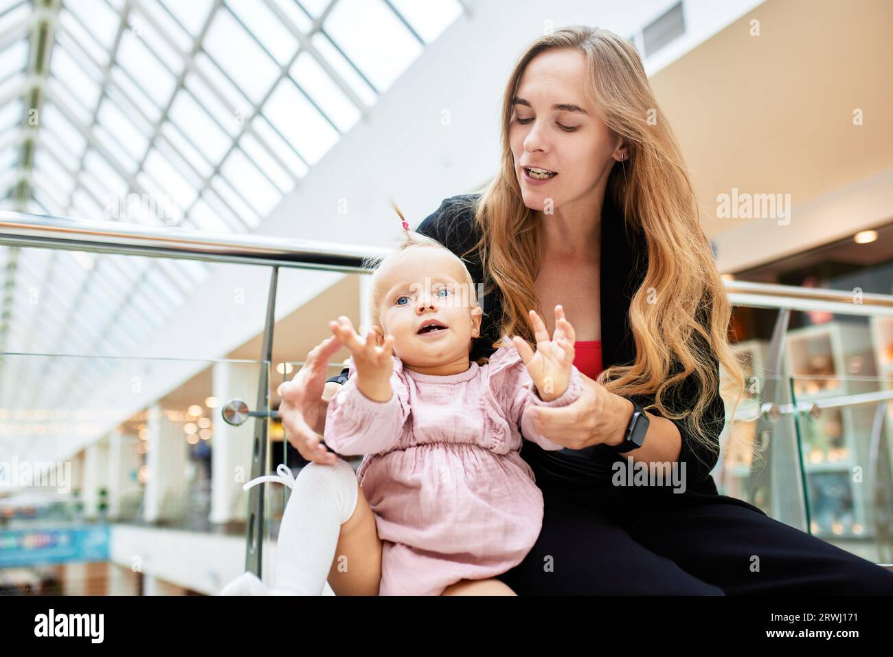 Jeune jolie mère avec un petit bébé assis sur un banc dans un centre commercial. Maman et fille dans les vêtements roses câliner, jouer, se détendre et s'amuser tout en sh Banque D'Images