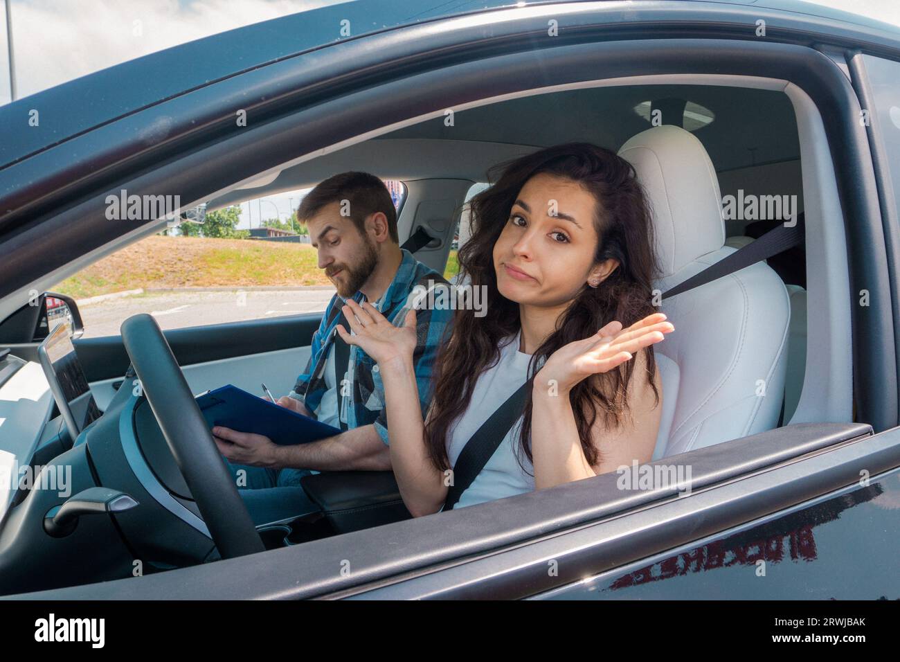 Jeune femme assise sur la place du conducteur et regardant la caméra avec confusion pendant que l'instructeur remplit le rapport d'essai routier. Test de conduite, conducteur Banque D'Images