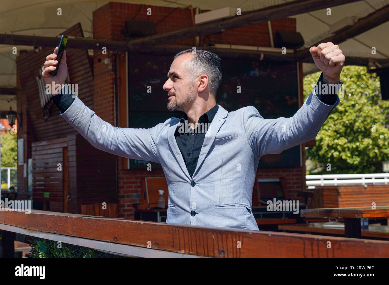 Homme d'affaires adulte caucasien avec veste, assis à l'extérieur du restaurant à midi en utilisant un téléphone portable avec les bras ouverts célébrant son succès et ses profits dans Banque D'Images