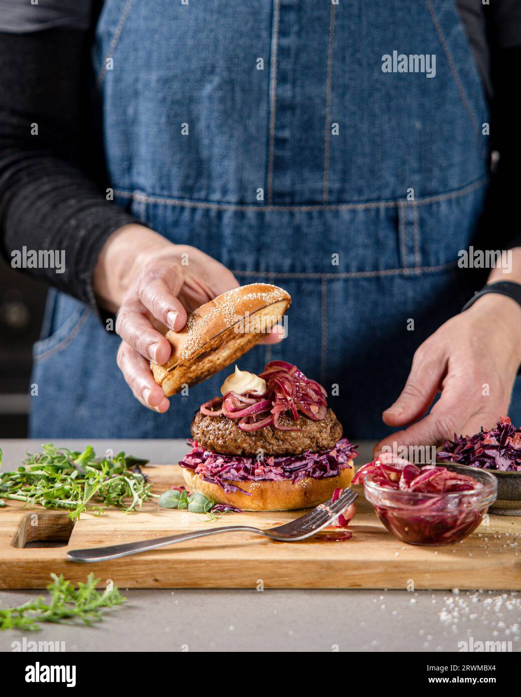 un hamburger maison prend le devant de la scène sur une planche à découper. La planche à découper est ornée de divers légumes et d'un petit bol d'oignons rouges marinés. Banque D'Images