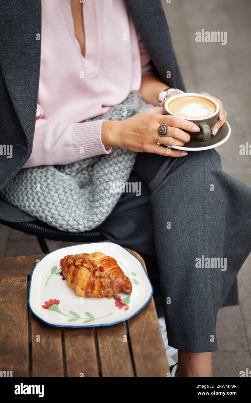 Fille est assise dans le café et tenant une tasse de café cappuccino dans ses mains, confortablement enveloppé dans une écharpe à carreaux gris. Gros plan des mains féminines. Banque D'Images