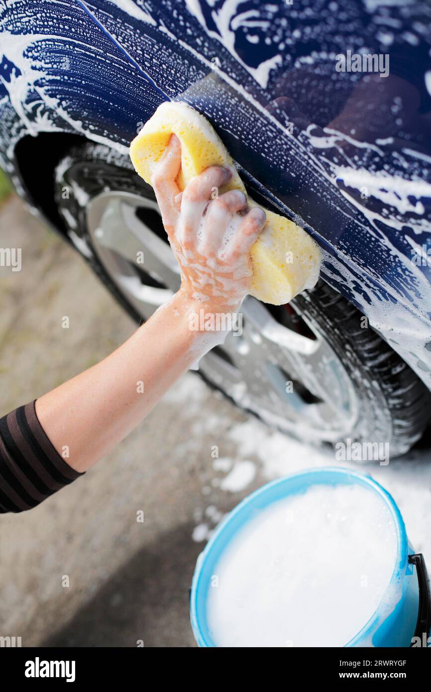 Homme lavant la voiture bleue avec une éponge jaune et un seau d'eau savonneuse Banque D'Images