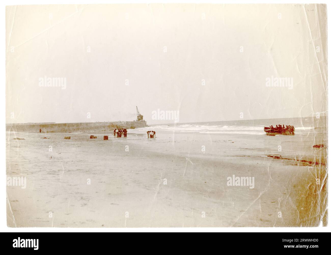 Vue de la plage de Jamestown, Accra, montrant le brise-lames avec une grande grue sur la jetée. Il y a plusieurs travailleurs africains qui traînent des marchandises sur la plage et un bateau à rames en bois est poussé sur la plage par un groupe d'hommes. Légende du manuscrit original : montrer le brise-lames à marée basse. Jamestown Accra. Banque D'Images