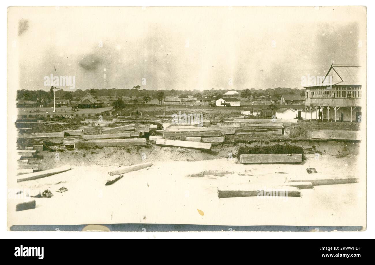 Vue d'un site industriel, peut-être Millers Ltd, avec de grands entrepôts et des blocs d'acajou se trouvant autour. Il y a un champ dans la distance avec poteau de drapeau et peut-être un pavillon. Banque D'Images
