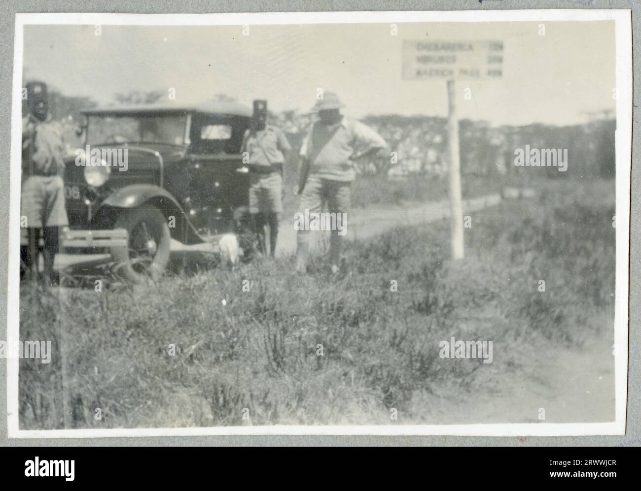 M. Bungey appuyé sur le côté d'une voiture noire garée sur le côté de la route. Derrière lui se trouve un poteau indicateur. Légende originale du manuscrit : sur la route dans le pays Turkana, en route pour Kapenguria. H.Q. pour S&W Turkana. Banque D'Images