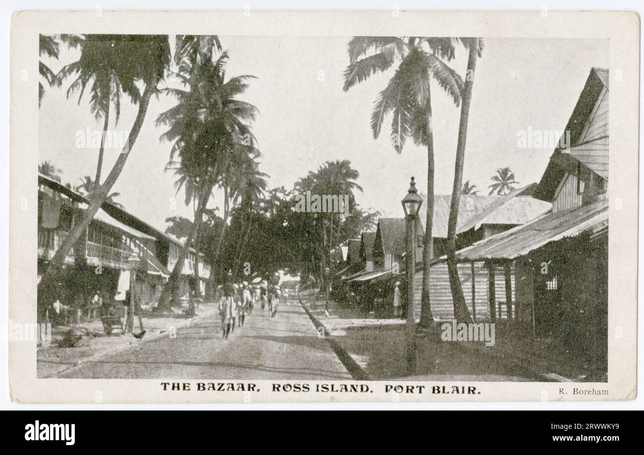 Carte postale montrant une rue bordée de palmiers, avec deux étages de bâtiments à ossature en bois de chaque côté. Les bâtiments ont des vérandas supérieures d'un côté et des pignons supérieurs de l'autre. Une ligne de piétons est représentée marchant le long du centre de la route. Décrit dans la légende imprimée comme le Bazaar, Ross Island, Port Blair. Légende au verso : magasins natifs. Ma véranda arrière regarde en bas de cette rue. Droit d'auteur : R Boreham. Banque D'Images