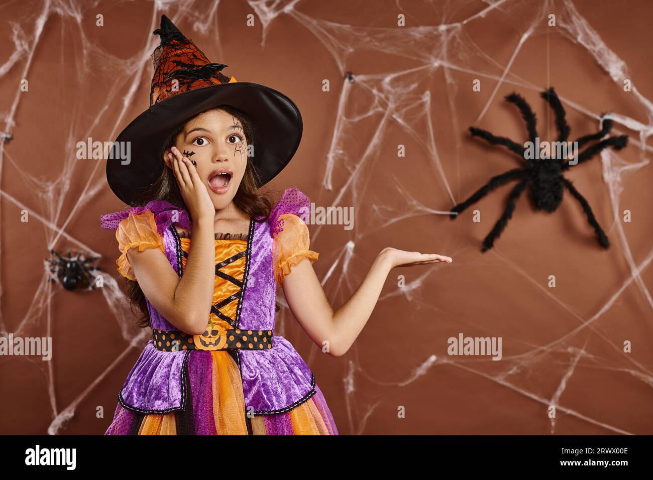 Fille émerveillée dans le chapeau de sorcière et costume d'Halloween pointant vers la fausse araignée sur fond brun, effrayante Banque D'Images