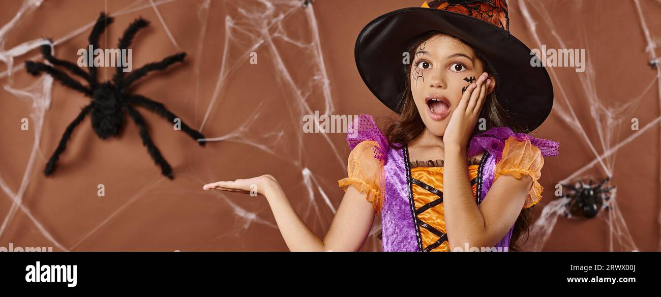 Fille émerveillée dans le chapeau de sorcière et costume d'Halloween pointant vers fausse araignée sur fond brun, bannière Banque D'Images