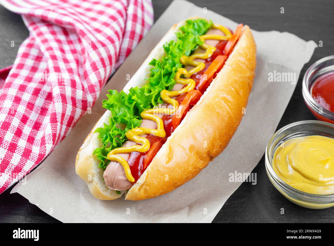 Hot Dog maison avec moutarde jaune, ketchup, tomate et feuilles de salade fraîches sur fond d'ardoise noire. Restauration rapide, cuisine de rue, cuisine américaine Banque D'Images