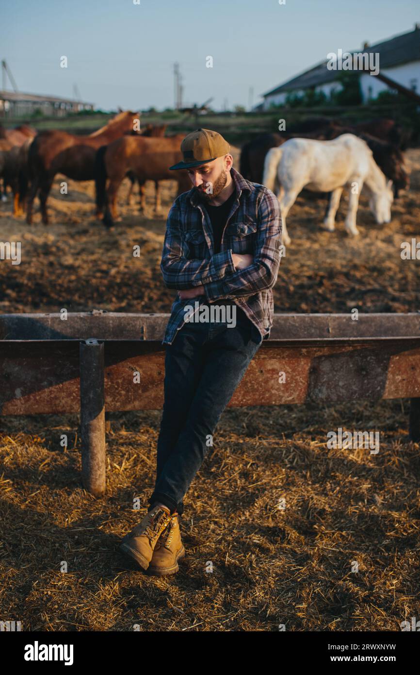 Portrait d'un jeune ouvrier agricole, sur le fond d'une clôture avec des chevaux. Un jeune agriculteur s’occupe des chevaux dans les écuries. Banque D'Images