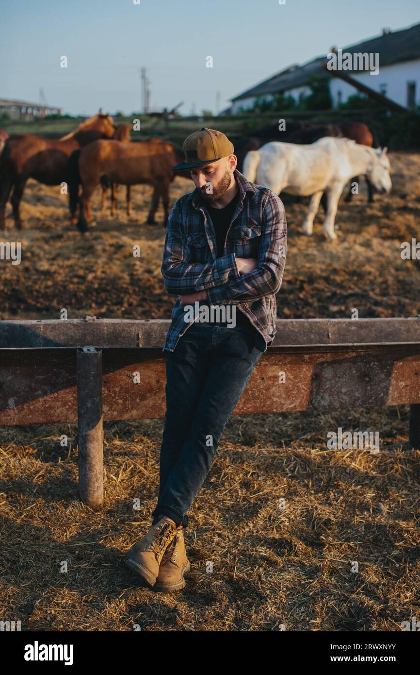 Portrait d'un jeune ouvrier agricole, sur le fond d'une clôture avec des chevaux. Un jeune agriculteur s’occupe des chevaux dans les écuries. Banque D'Images