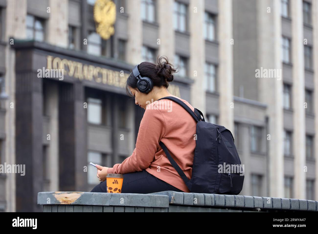 Fille dans les écouteurs assis avec un smartphone et boire un café sur le banc contre la Douma d'État à Moscou Banque D'Images