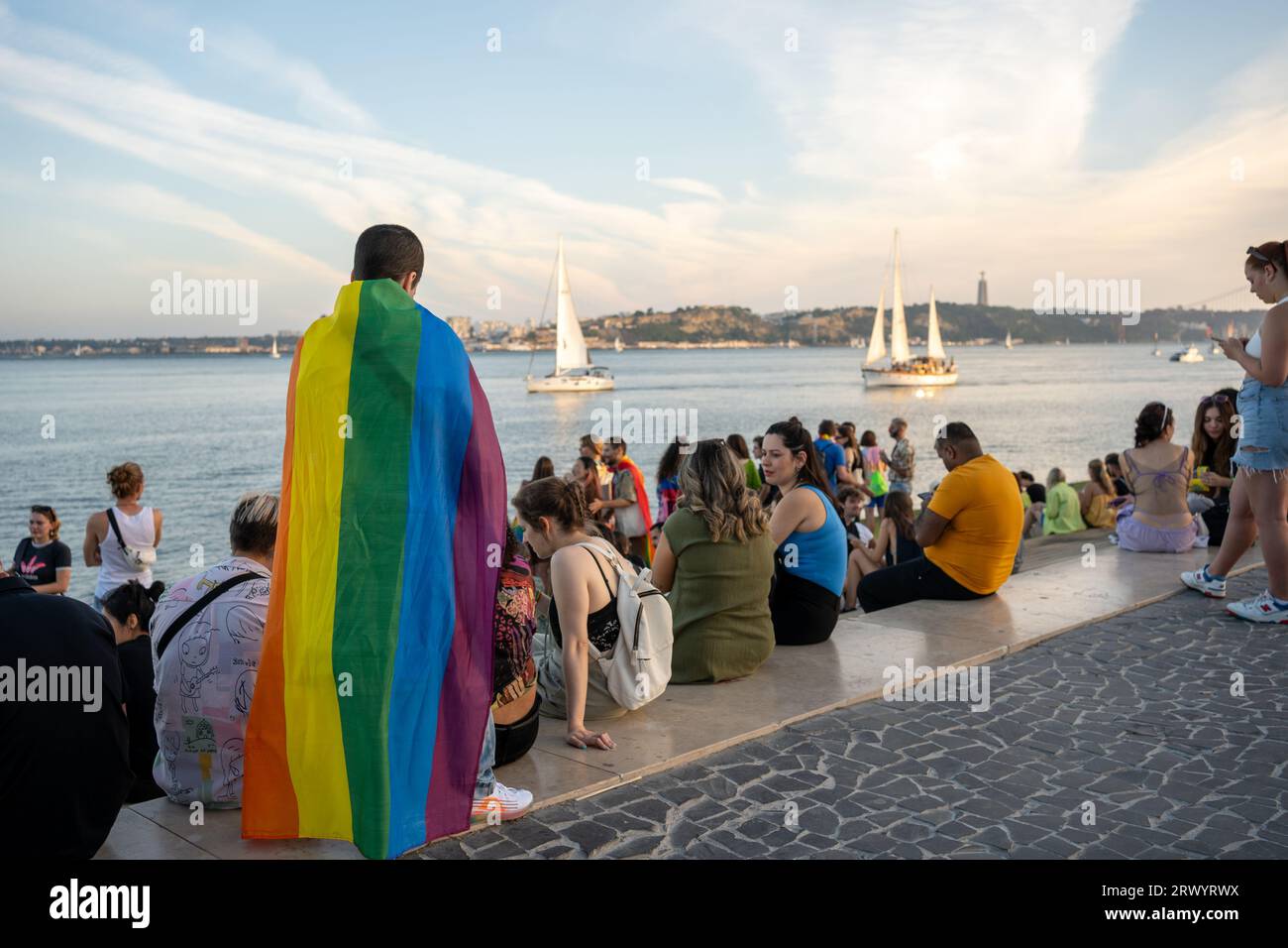 Lisbonne, Portugal. 17 juin 2023 Homme avec drapeau arc-en-ciel gay défilé à promenade. Homme gay debout à l'extérieur pendant le mois de la fierté. Communauté LGBT portant un drapeau arc-en-ciel regardant un yacht sur l'eau Banque D'Images