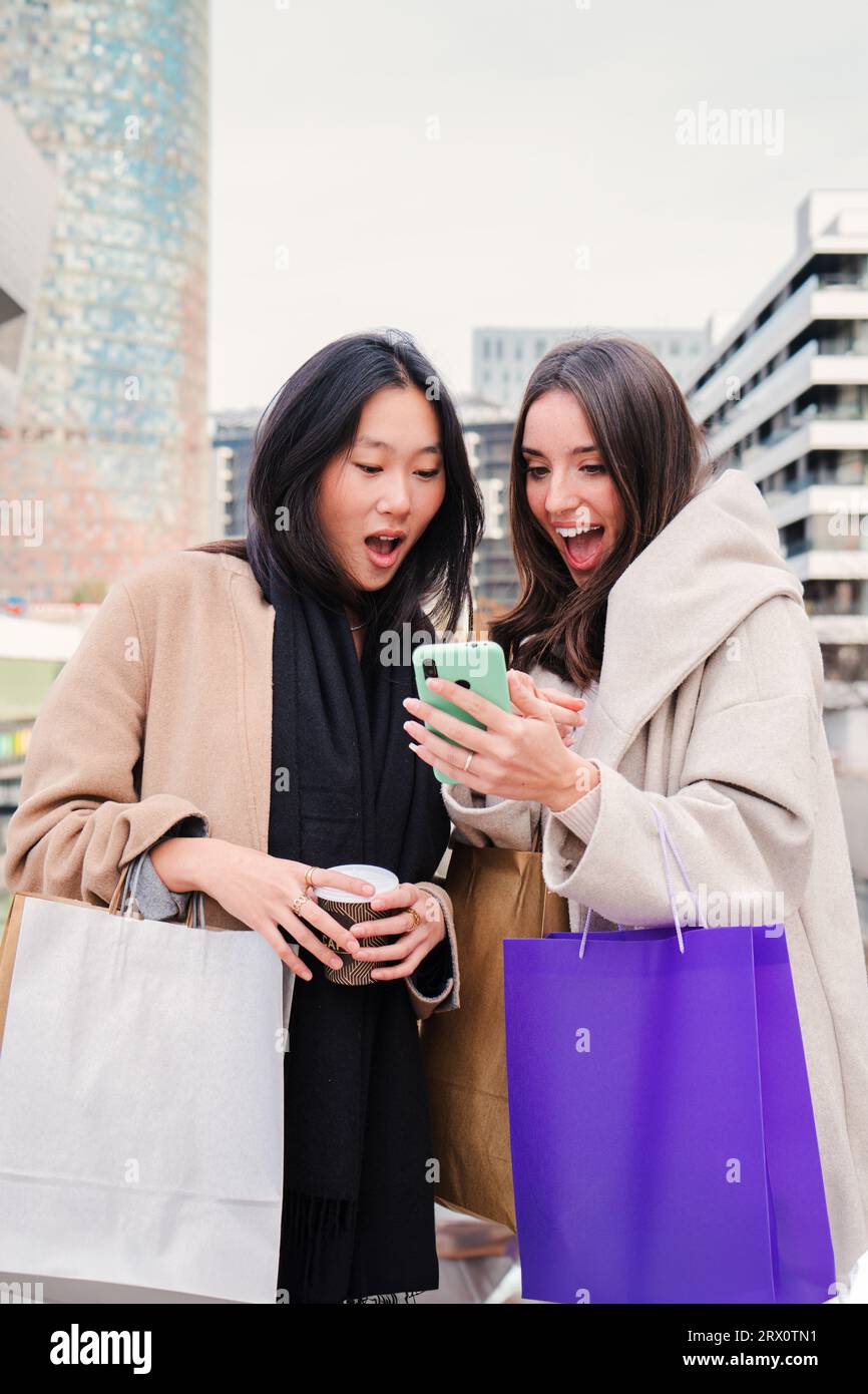 Portrait vertical de jeunes femmes heureuses multiethniques surpris par les ventes et les offerts à la recherche d'un téléphone portable faisant des achats en ligne. Amies consommatrices achetant des vêtements à l'aide d'un smartphone. Photo de haute qualité Banque D'Images