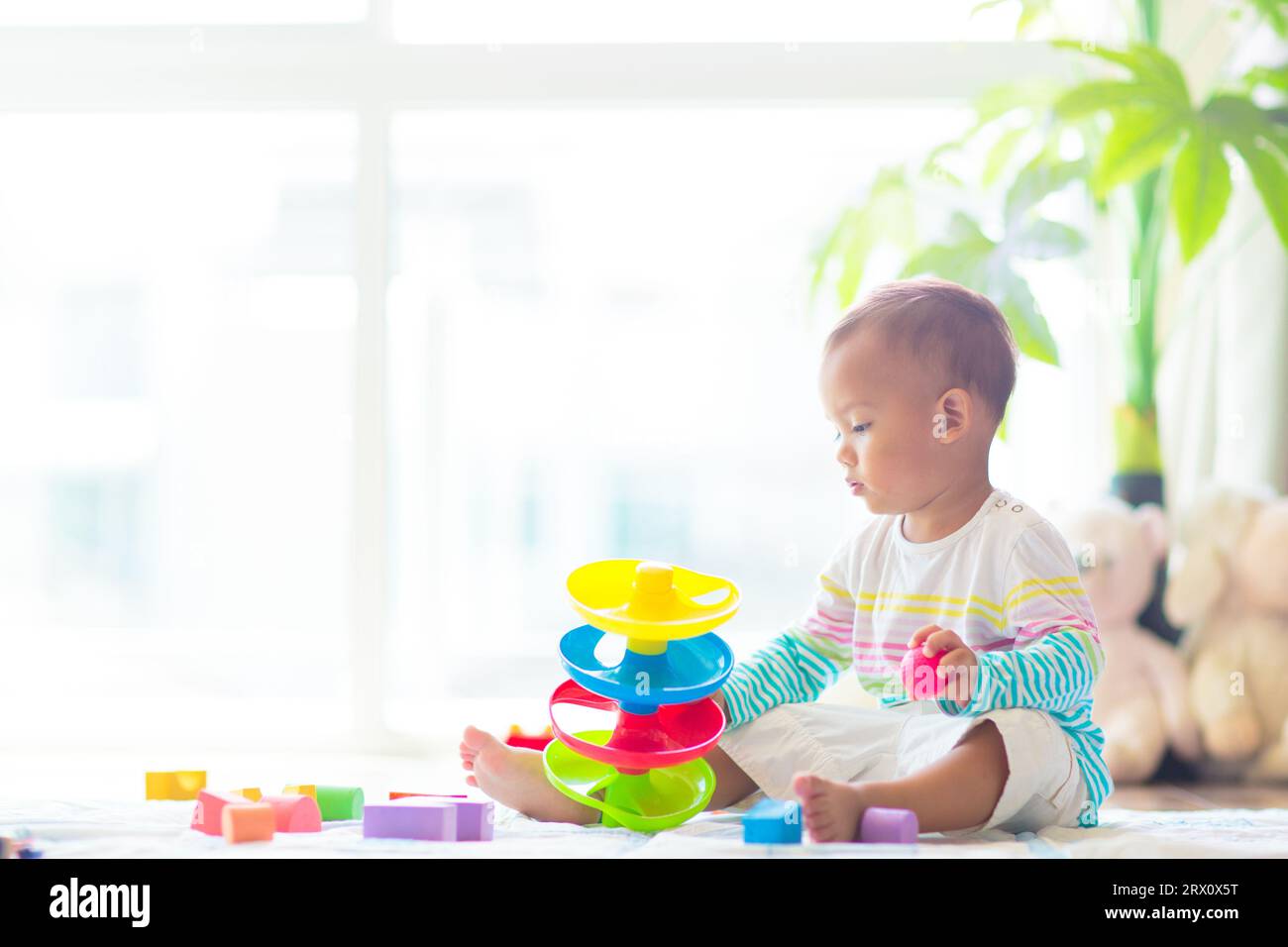 Adorable bébé asiatique apprenant à ramper et à jouer avec le jouet coloré de bloc dans la chambre blanche ensoleillée. Mignon rire enfant rampant sur un tapis de jeu. Banque D'Images