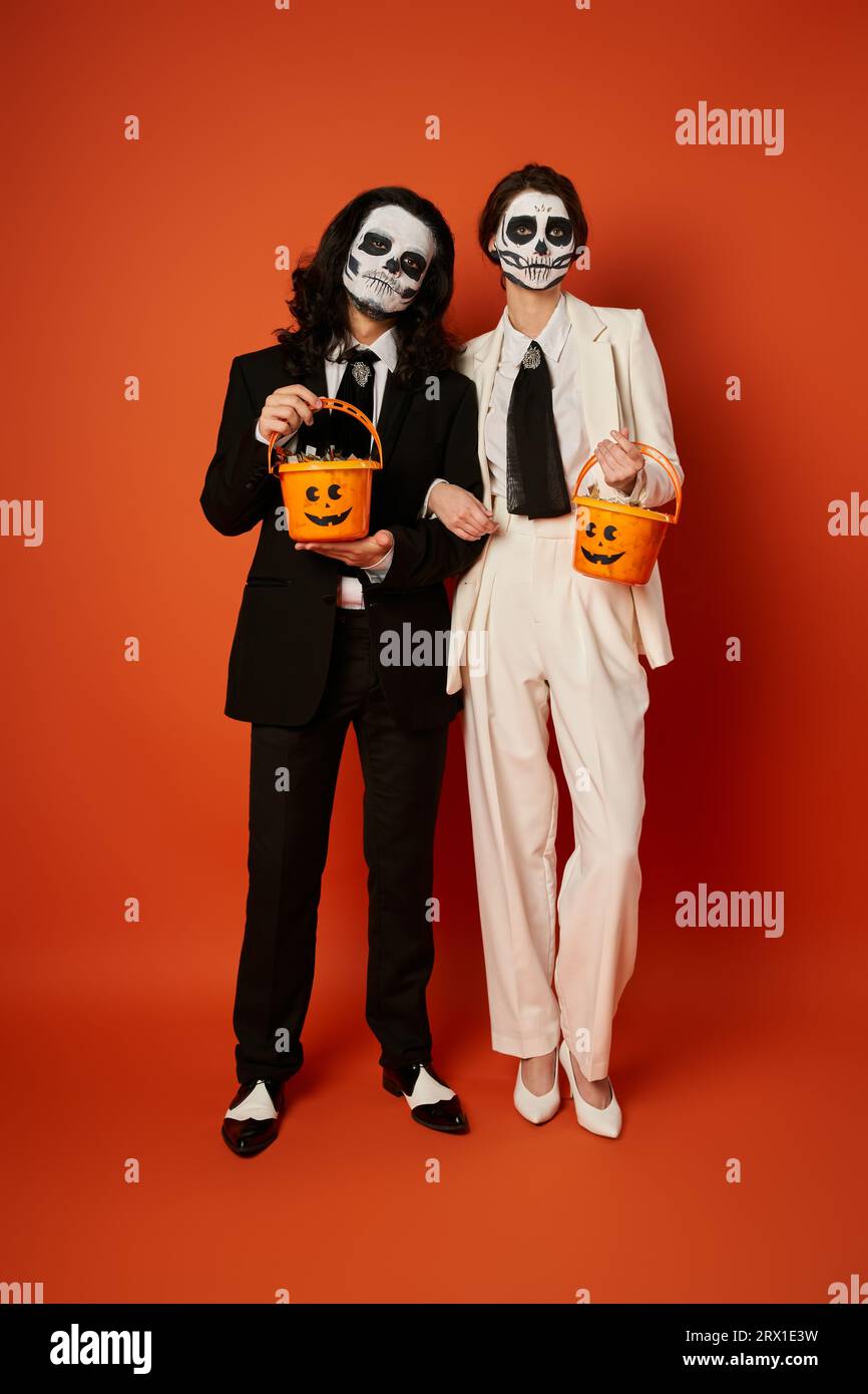 Toute la longueur d'un couple élégant en maquillage catrina tenant des seaux de bonbons sur le rouge, Day of Dead fest Banque D'Images