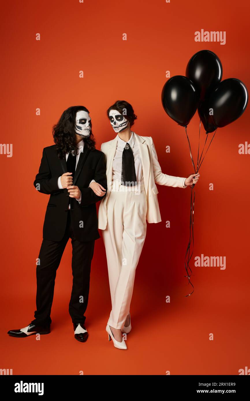 Couple en maquillage catrina et costumes se regardant près de ballons noirs sur rouge, Day of Dead Banque D'Images