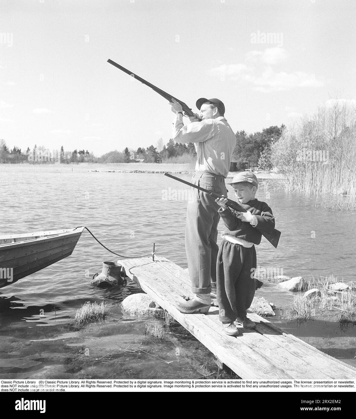 Père et fils dans les années 1950 Ils se tiennent debout avec leurs fusils sur un quai près d'un lac avec leurs fusils de chasse prêts. Suède 1956. Kristoffersson réf. BX77-12 Banque D'Images