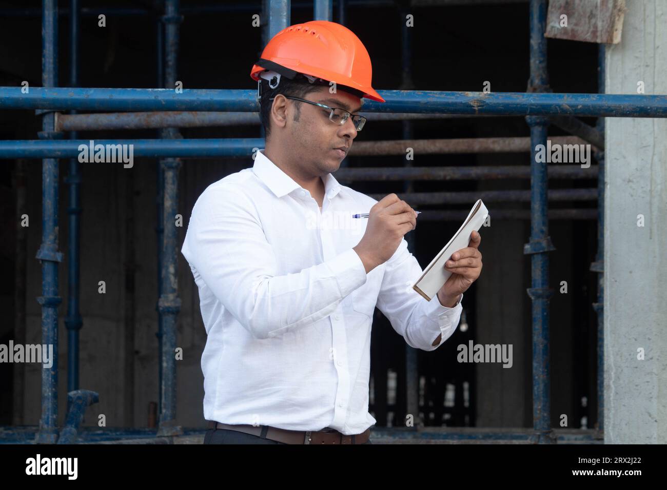 Ingénieur masculin en chemise blanche et en casque dur écrivant des notes sur le chantier de construction. Il est concentré tenant le bloc-notes et le stylo Banque D'Images