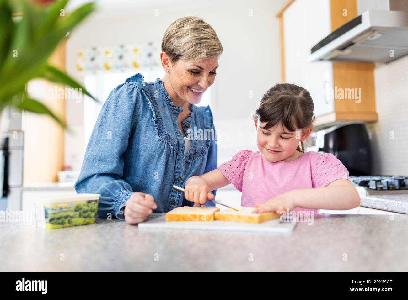 Fille étalant le beurre sur le pain par mère debout dans la cuisine Banque D'Images