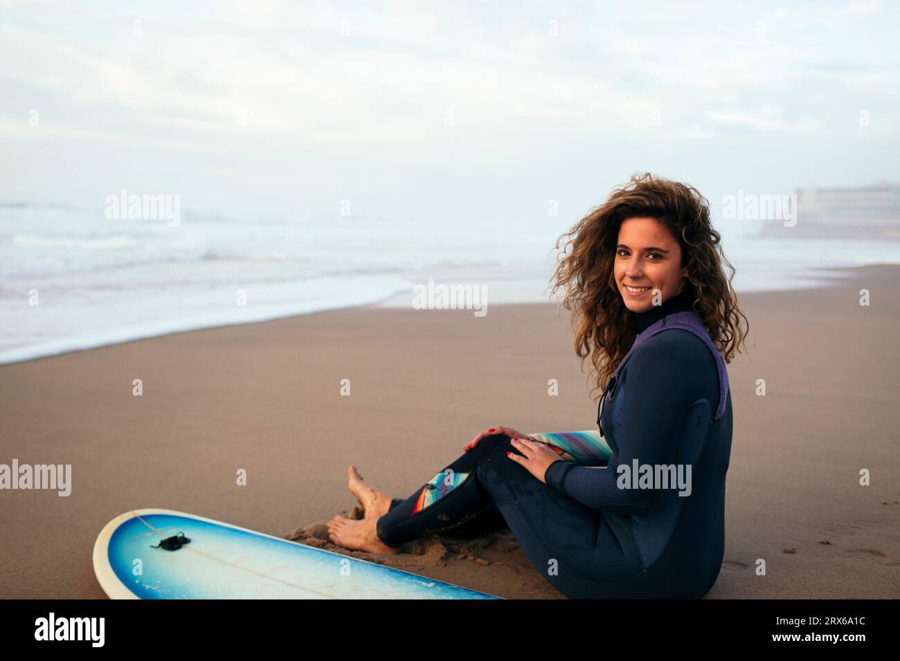 Femme souriante assise avec planche de surf à la plage en vacances Banque D'Images