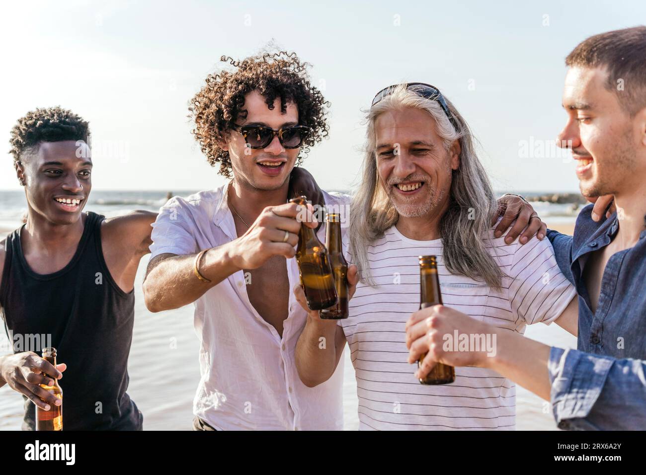 Amis souriants grillant des bouteilles de bière à la plage par jour ensoleillé Banque D'Images