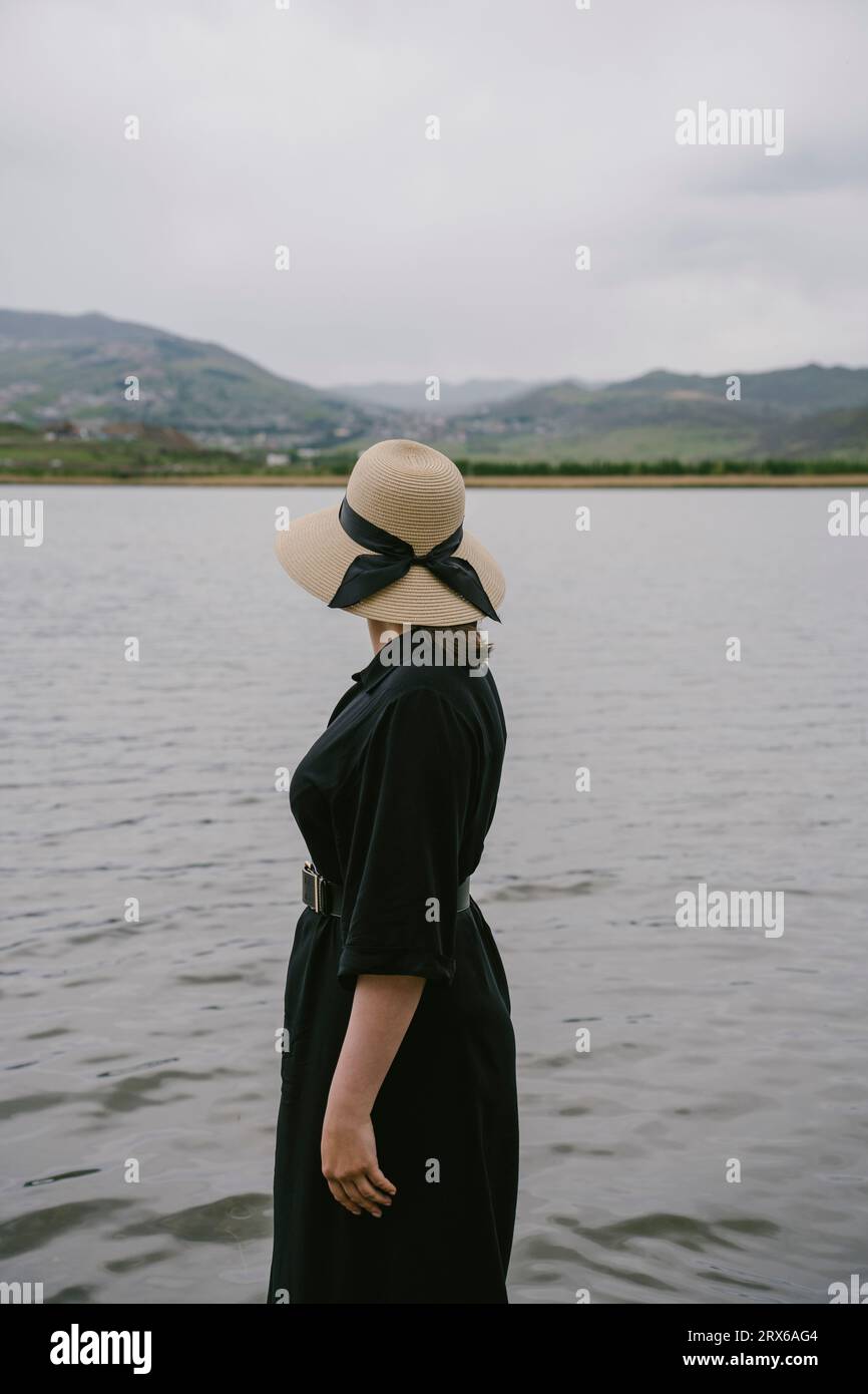 Femme réfléchie portant robe noire et chapeau de paille debout près du lac Banque D'Images