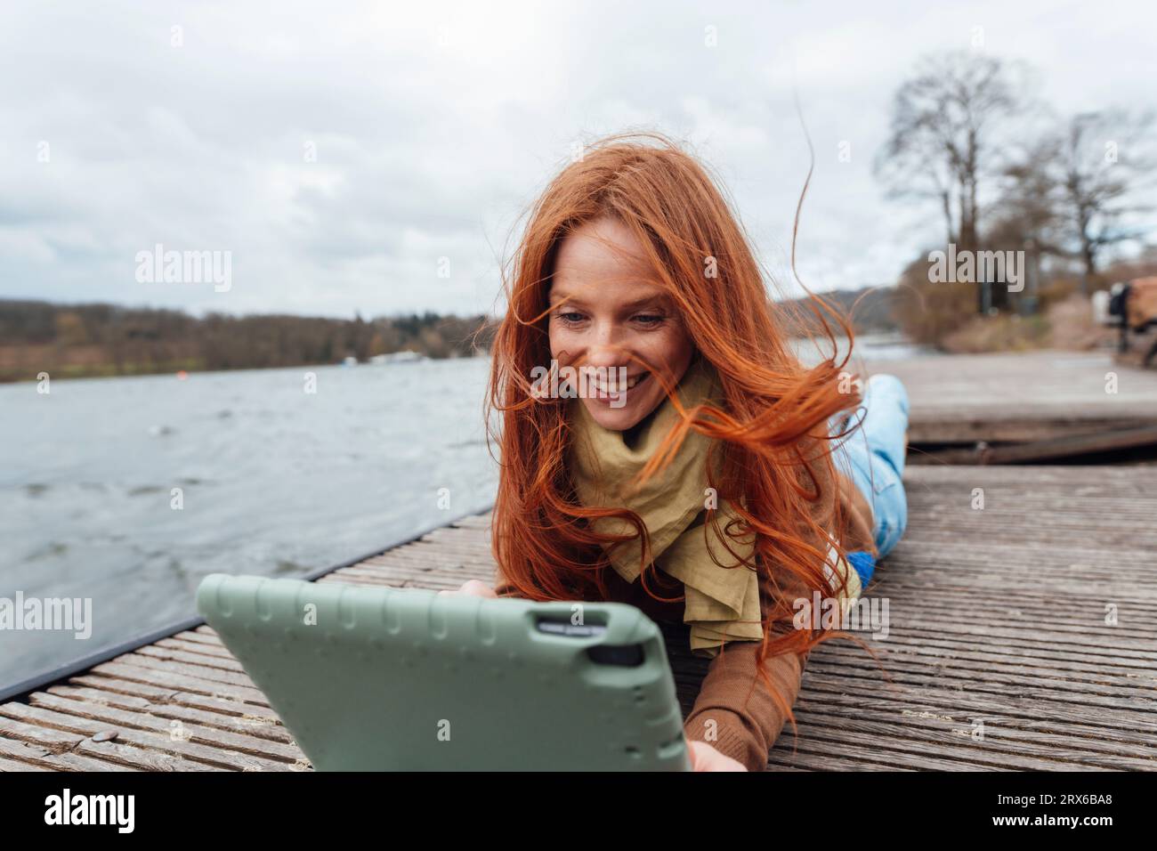 Femme rousse souriante à l'aide d'une tablette PC sur la jetée par le lac Banque D'Images