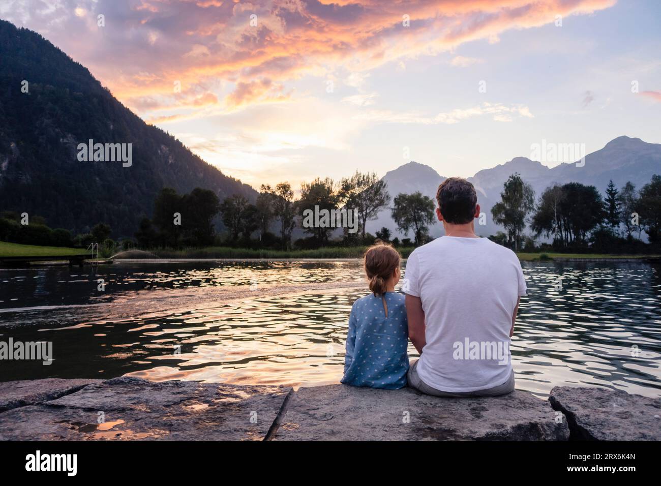 Père et fille assis sur le rocher et admirant le lac au coucher du soleil Banque D'Images