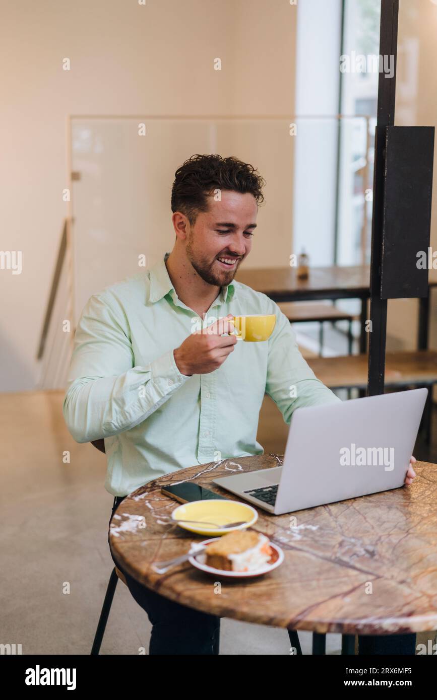 Homme d'affaires heureux avec tasse à café travaillant sur ordinateur portable à table dans le café Banque D'Images