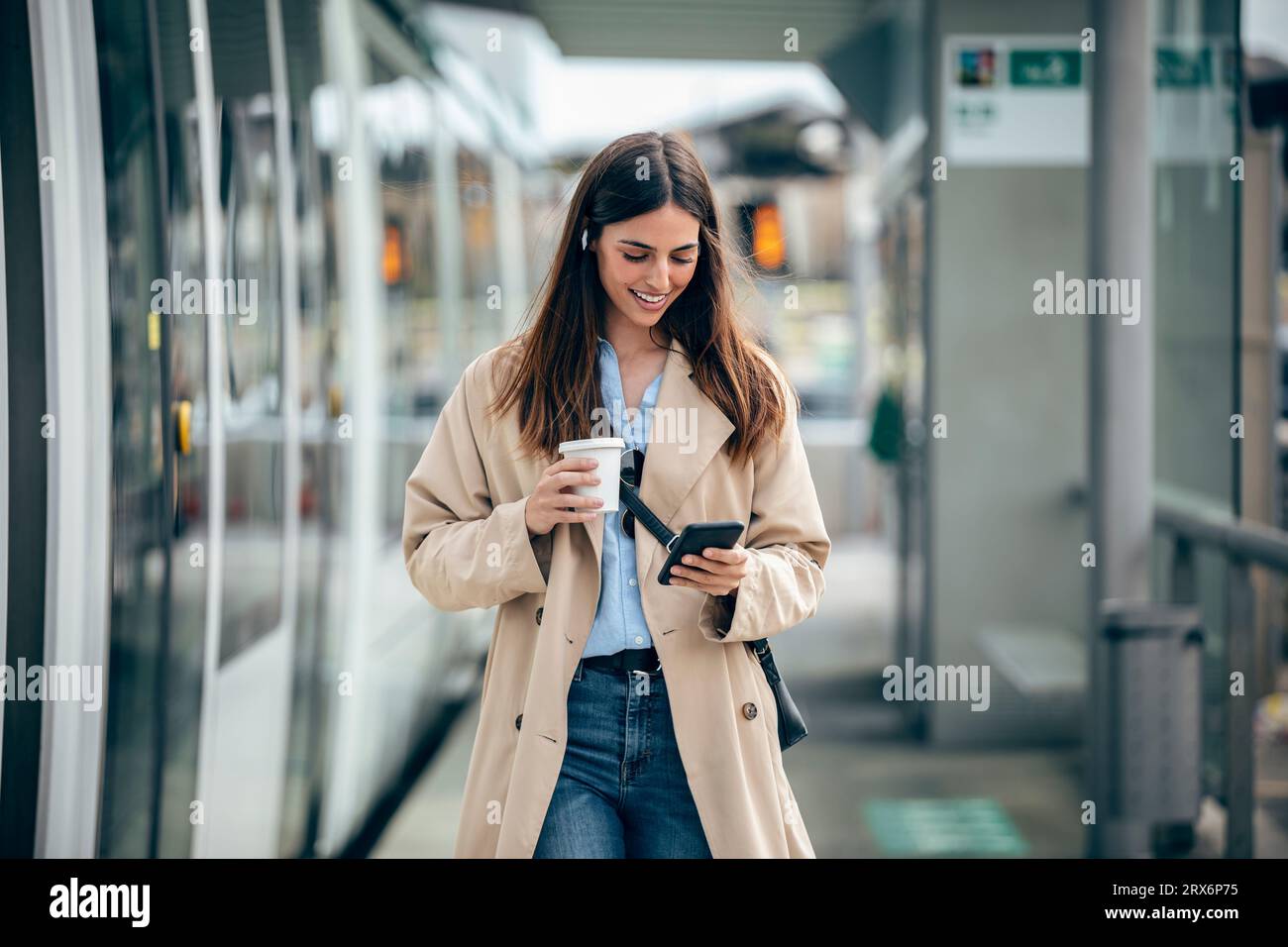 Femme heureuse marchant avec une tasse à café et en utilisant un téléphone intelligent à la station de tram Banque D'Images