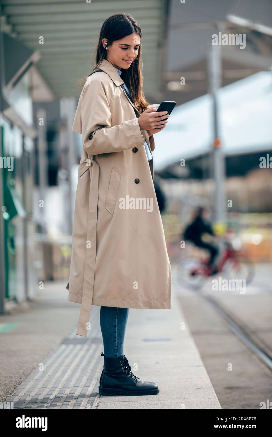 Femme souriante à l'aide d'un téléphone intelligent à la station de tram Banque D'Images