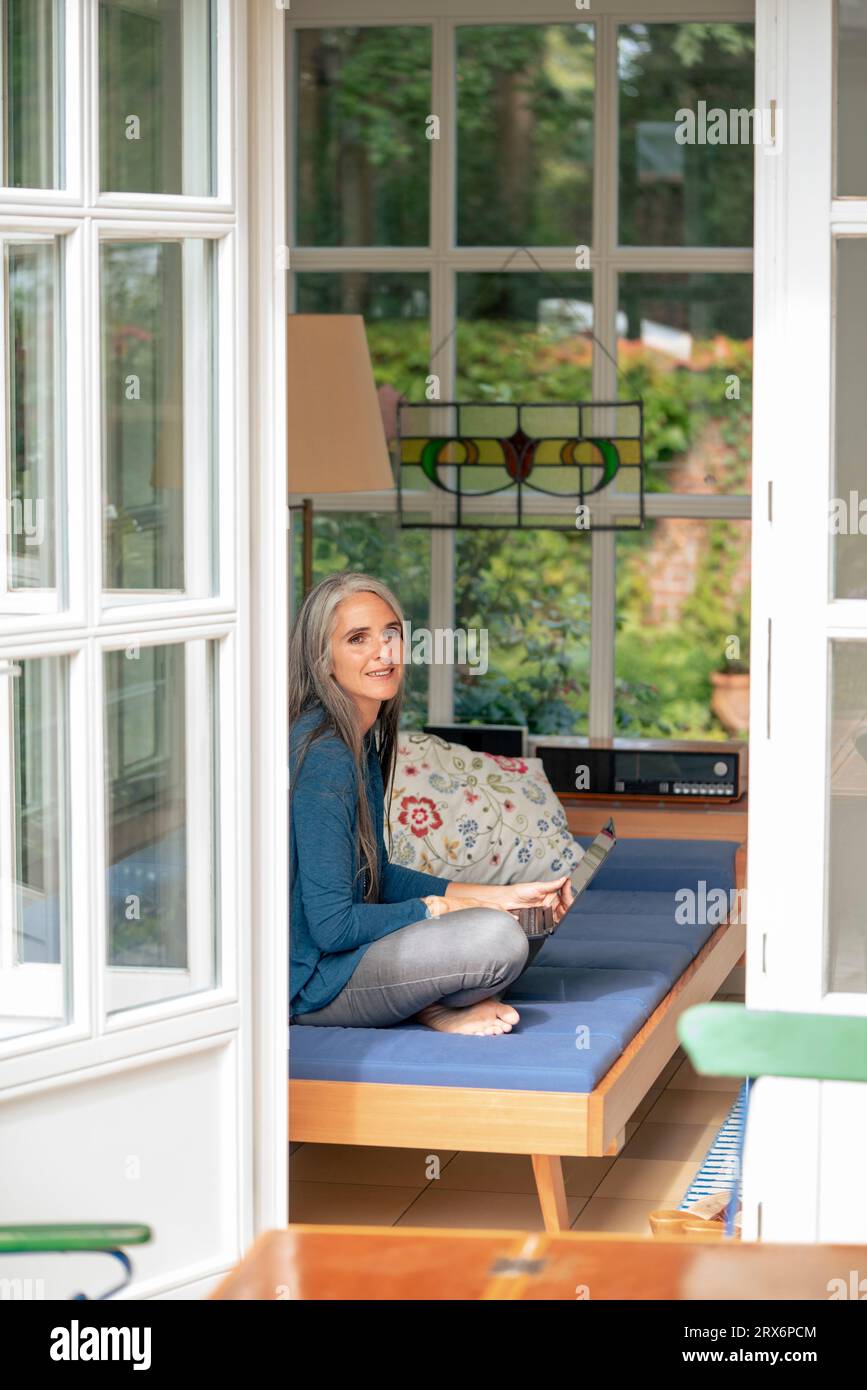Femme mature souriante assise avec ordinateur portable sur le canapé à la maison Banque D'Images