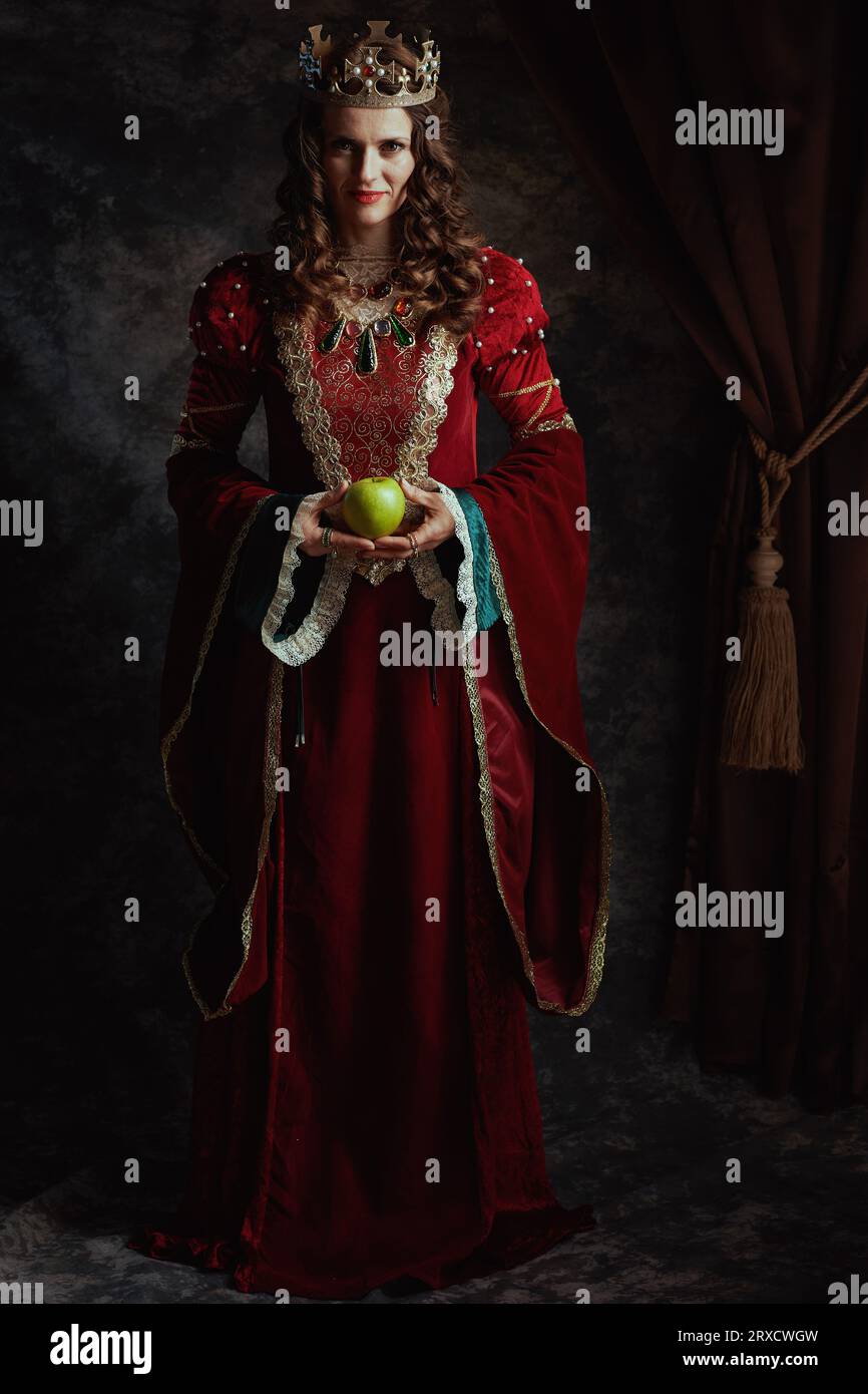 Portrait en longueur de reine médiévale en robe rouge avec pomme verte et couronne sur fond gris foncé. Banque D'Images