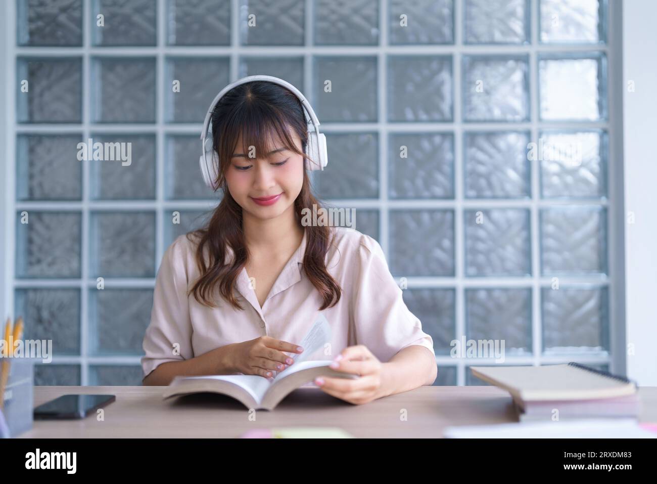 Jeune étudiante asiatique en vêtements décontractés lisant des manuels scolaires pour étudier les connaissances en éducation et faire des devoirs tout en portant des écouteurs et des lis Banque D'Images