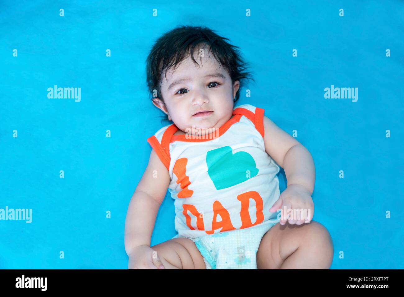 Six mois mignon petit garçon indien jouer en portant i love papa t-shirt isolé sur fond de studio bleu, enfant heureux avec les cheveux noirs. Banque D'Images