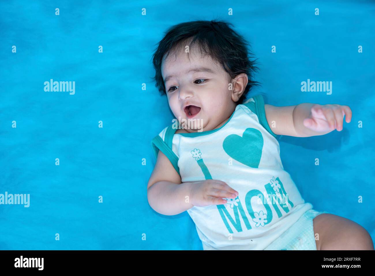 Six mois mignon petit garçon indien jouer en portant i love mom t-shirt isolé sur fond de studio bleu, enfant heureux avec les cheveux noirs. Banque D'Images