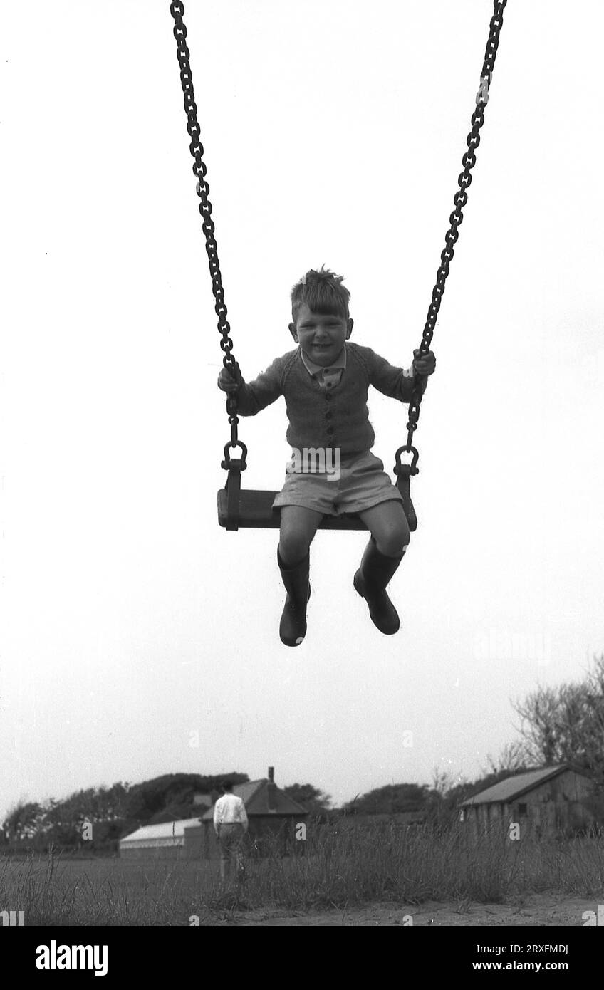 Années 1960, historique, un jeune garçon dans les airs, chevauchant une balançoire taditionnelle, tenant sur la chaîne de balançoire en métal, Angleterre, Royaume-Uni. Banque D'Images