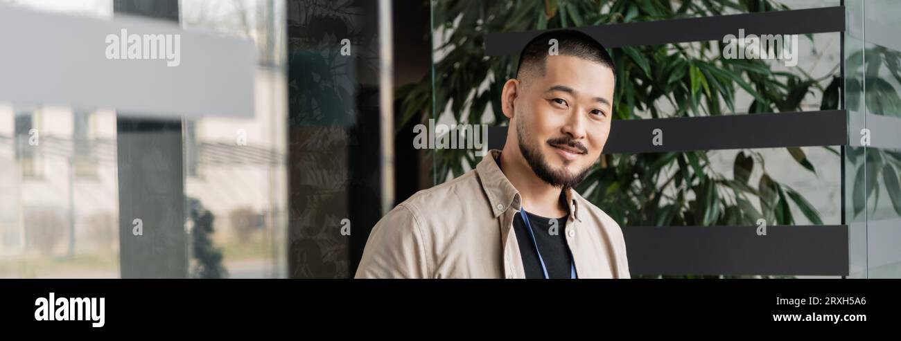 heureux homme d'affaires asiatique souriant et regardant la caméra près de la porte vitrée dans le bureau moderne, bannière Banque D'Images